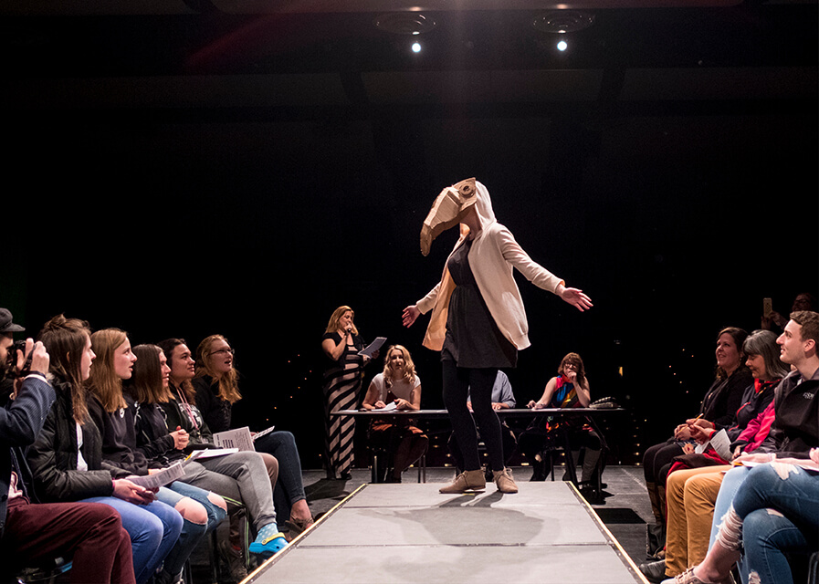 Student walking on stage showing a mask they made in an art course.