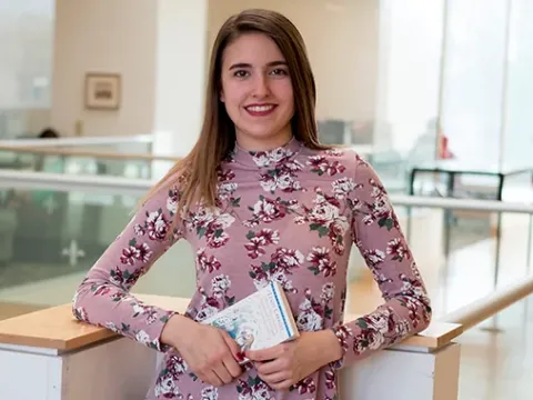 Female student stands in a library holding a book.