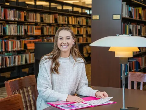 Female student sits at a table in the library with study materials.