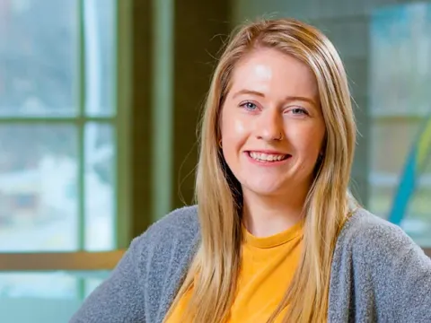 Female student posing in front of windows inside a building.